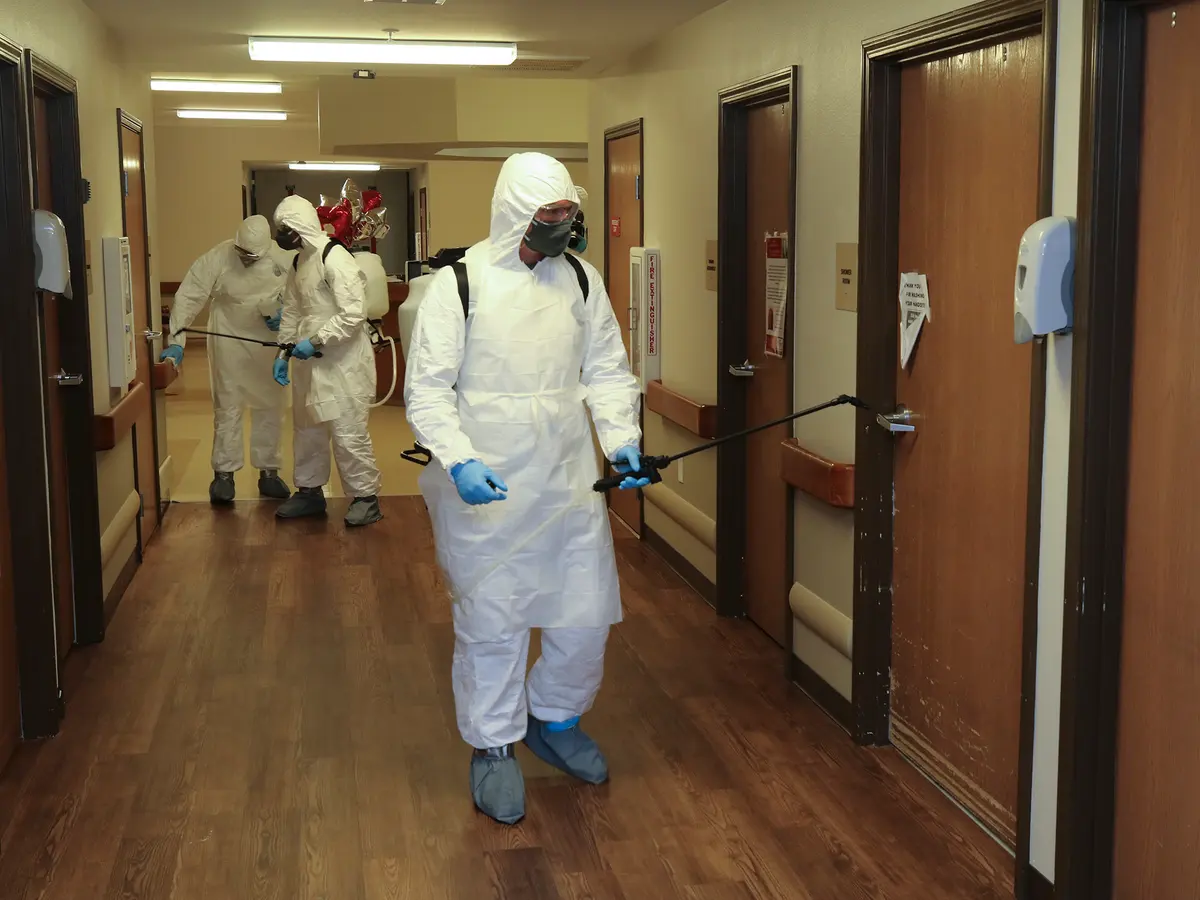 Técnicos con traje blanco desechable, capucha, mascarilla, antiparras y guantes usan pulverizadores de espalda junto a las puertas de un pasillo con pasamanos