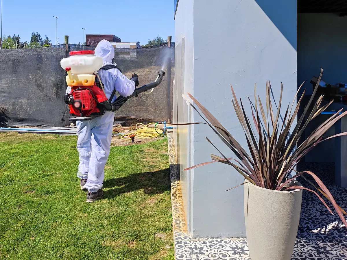 Técnico de VIS con buzo blanco aplicando con atomizadora motorizada en el cerco perimetral del patio de una casa