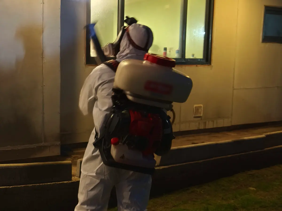 De noche, técnico de VIS con traje y atomizadora motorizada aplicando en el muro exterior junto a una ventana iluminada