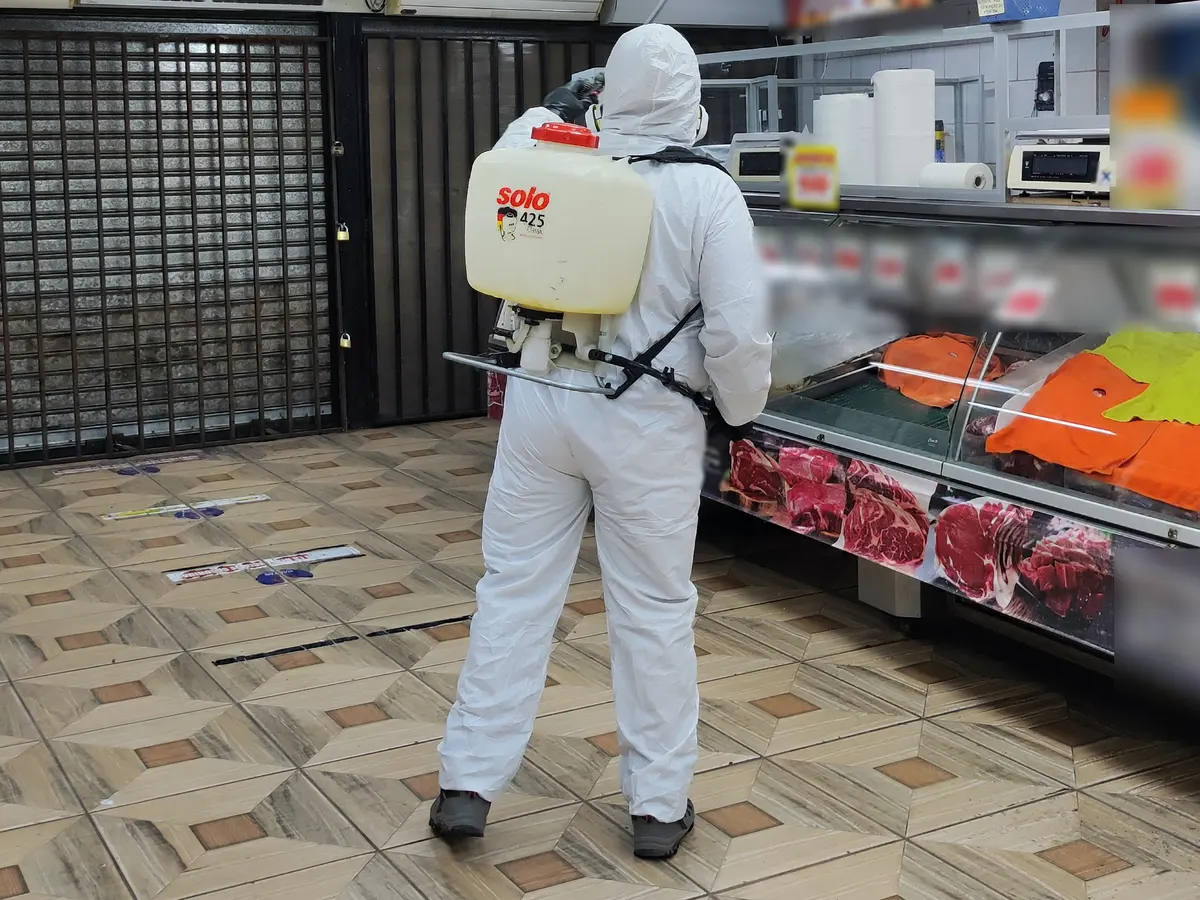 Técnico con traje de protección y bomba de espalda sanitizando un local de venta de carnes con la vitrina cubierta