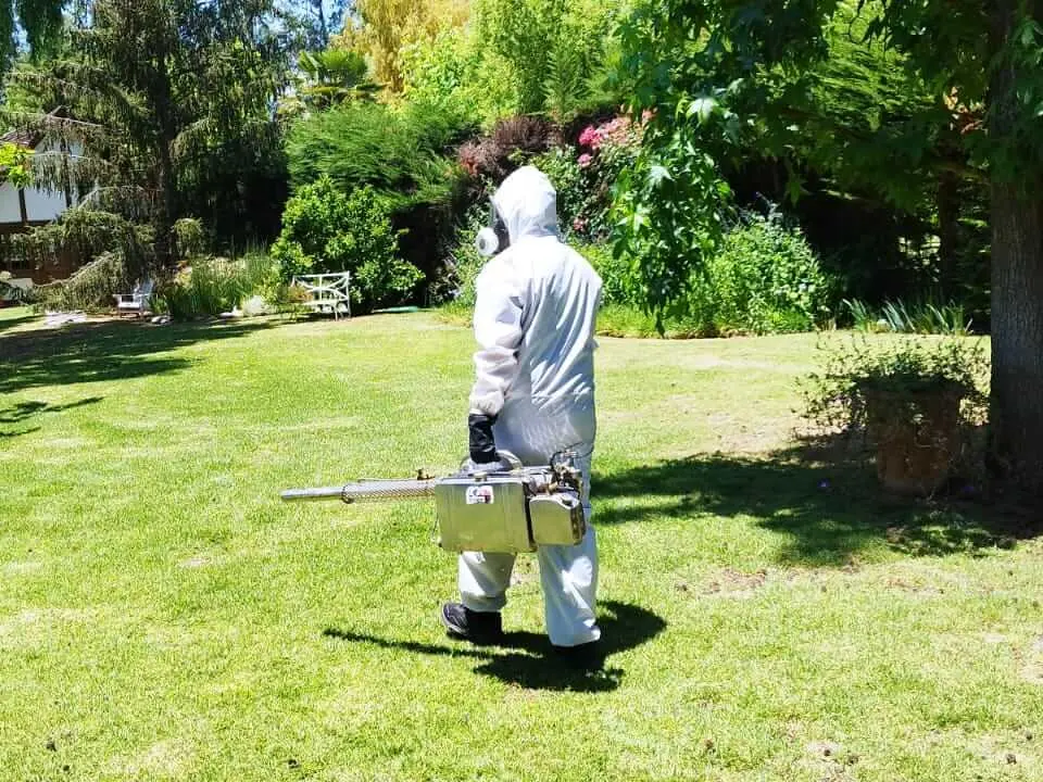Técnico con traje de protección y respirador caminando con un termonebulizador por un jardín con césped y árboles