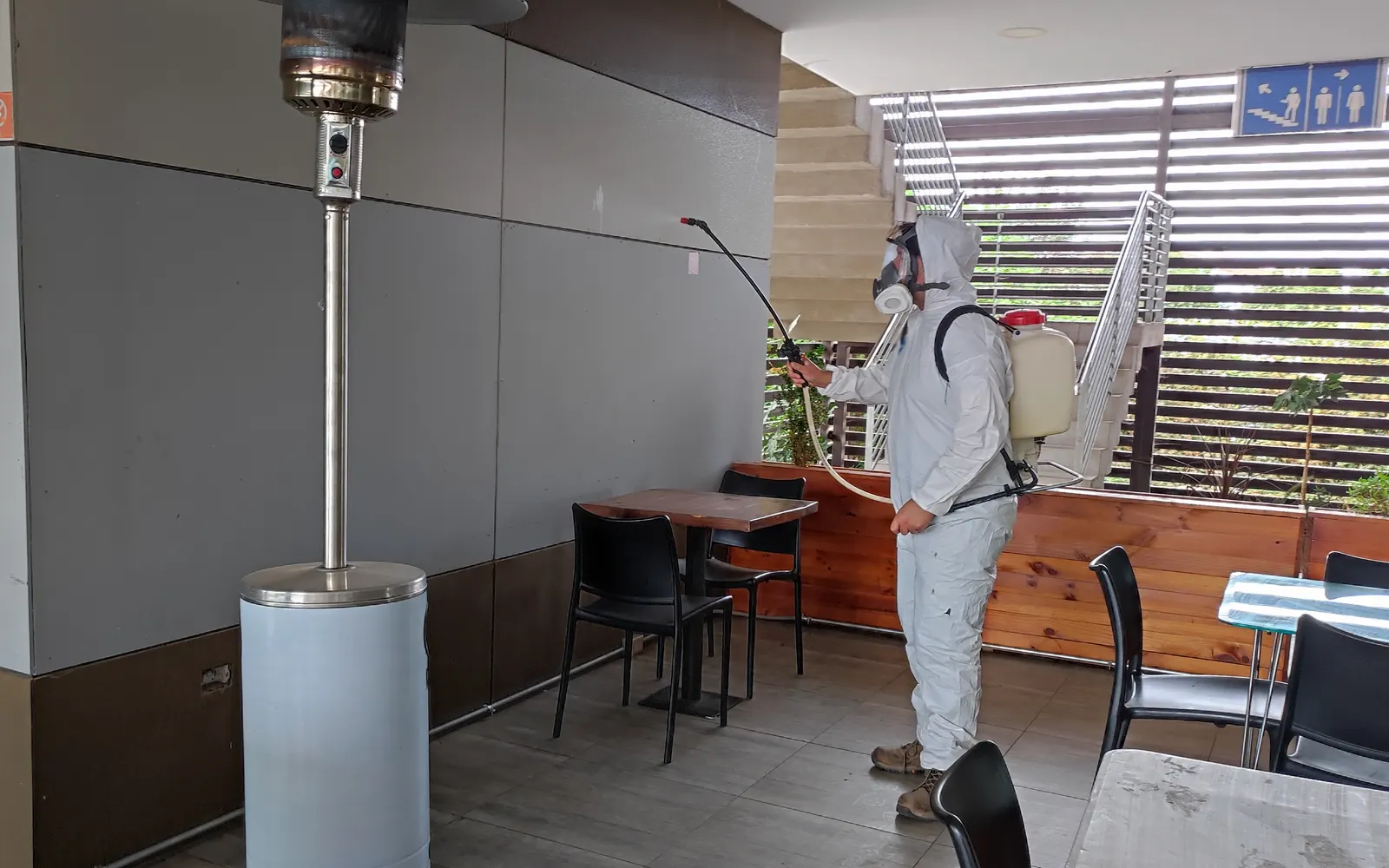 Técnico de VIS Control de Plagas con traje y máscara de rostro completo aplicando con pulverizador de espalda en la terraza de un restaurante
