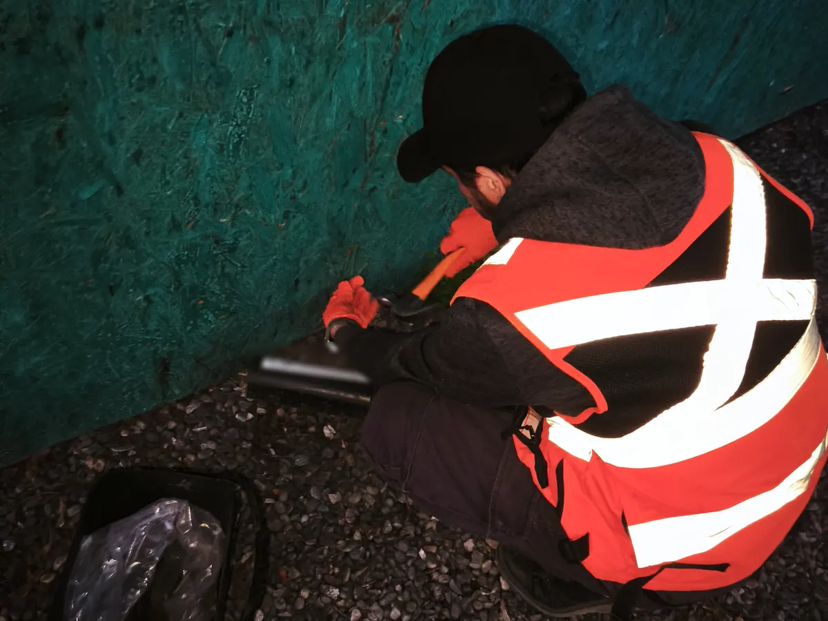 Técnico de VIS con chaleco reflectante y guantes fijando una estación cebadera tubular al pie de un cerco, sobre gravilla, durante una desratización perimetral