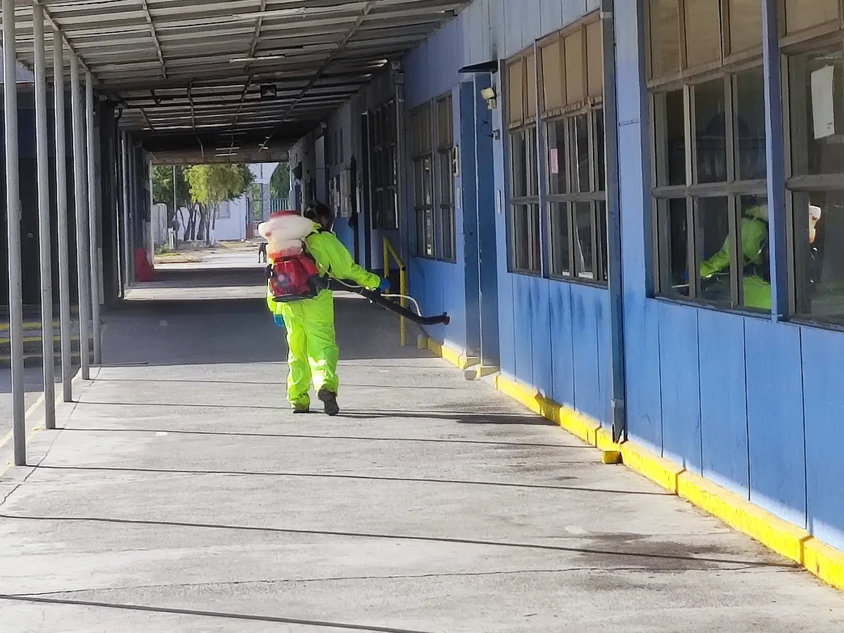 Técnico con traje amarillo flúor aplicando con atomizadora en un pasillo techado junto a salas, con otro técnico adentro